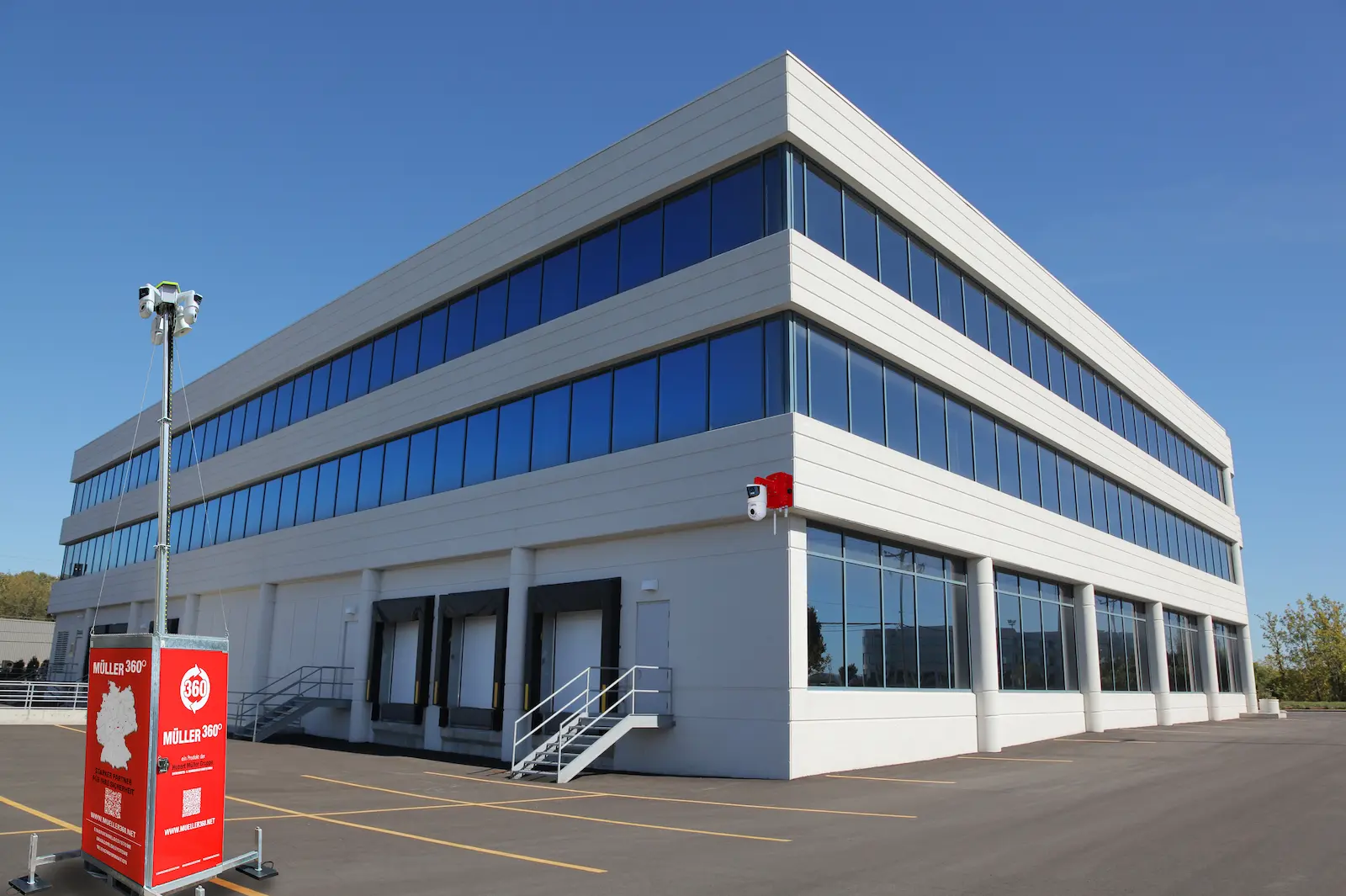 Moderner Gewerbe- oder Industriebau mit großflächigen Fensterfronten; davor ein roter Müller360-Videoturm der Hubert Müller Gruppe.