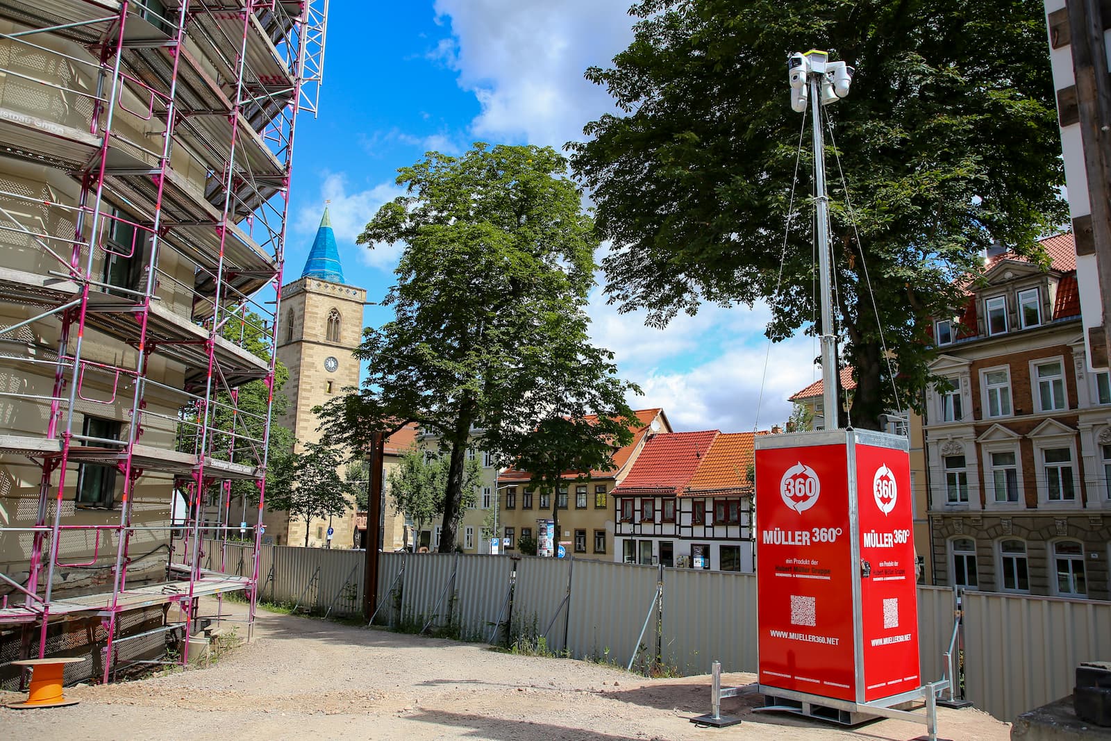 Roter Müller360-Videoturm der Hubert Müller Gruppe auf einer innerstädtischen Baustelle neben einem eingerüsteten Gebäude; im Hintergrund sind Bäume, Wohnhäuser und ein Kirchturm zu sehen.