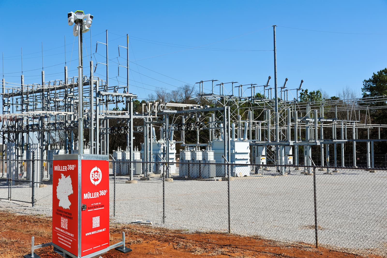 Elektrisches Umspannwerk mit technischen Anlagen und Leitungsstrukturen hinter einem Zaun; davor ein roter Müller360-Videoturm der Hubert Müller Gruppe.